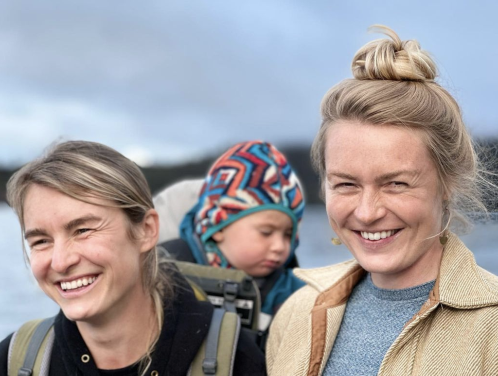 Two smiling blonde women, one with a baby on her back